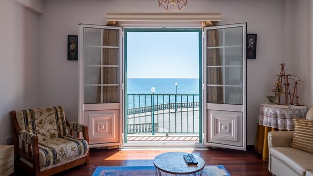 Ein Zimmer mit Meerblick, ein Holzstuhl, eine Couch, ein Tisch mit Fernbedienung und eine Zierpflanze.