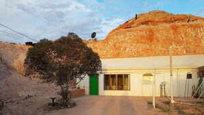 Un edificio con puerta verde, camino de tierra que lo conduce y una gran formación rocosa al fondo.