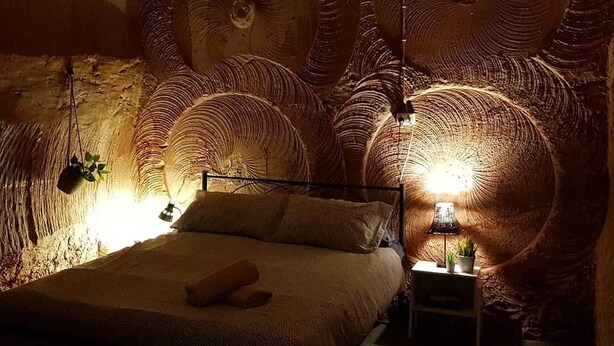 Una habitación circular y acogedora, con una cama ubicada en el centro, rodeada de paredes con textura y una lámpara colgante.