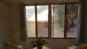 Habitación con una mesa, dos sillas, una planta en maceta y vistas a un paisaje desértico al atardecer a través de amplias ventanas.