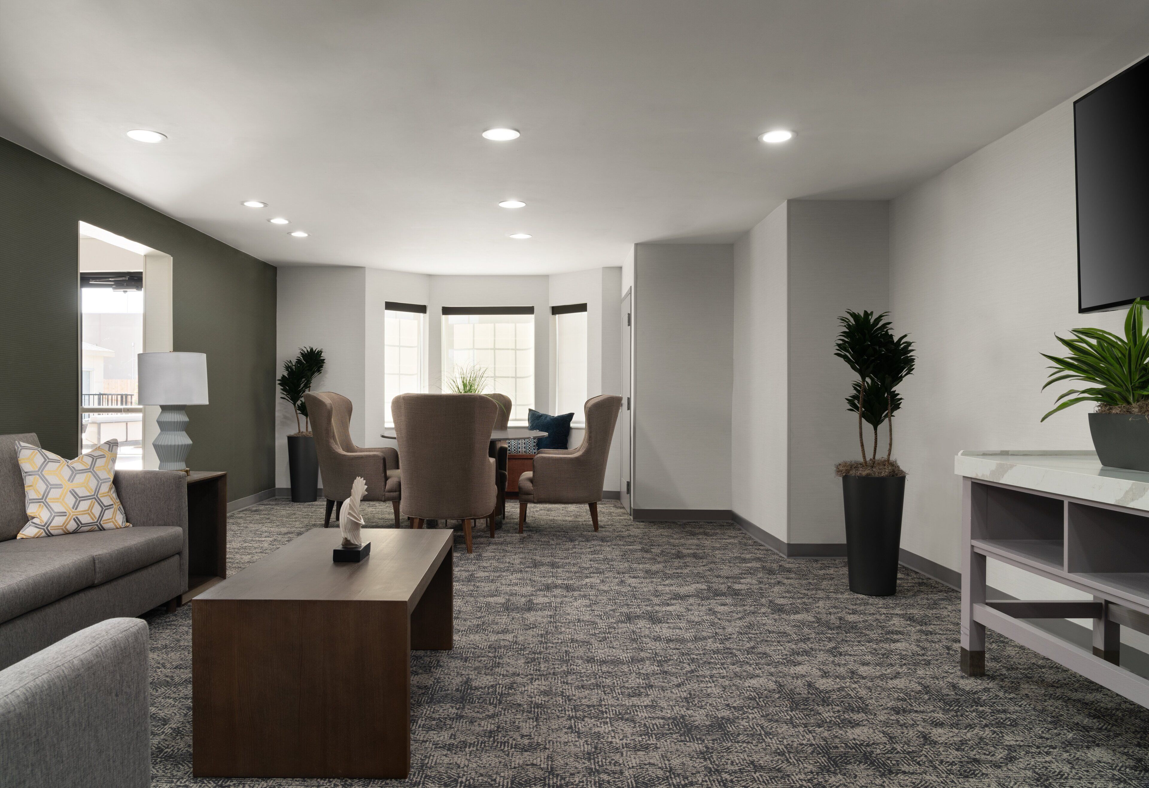 A modern living room with a grey sofa, a wooden coffee table, and a dining area with chairs and a table. There are potted plants and a flat-screen TV mounted on the wall.