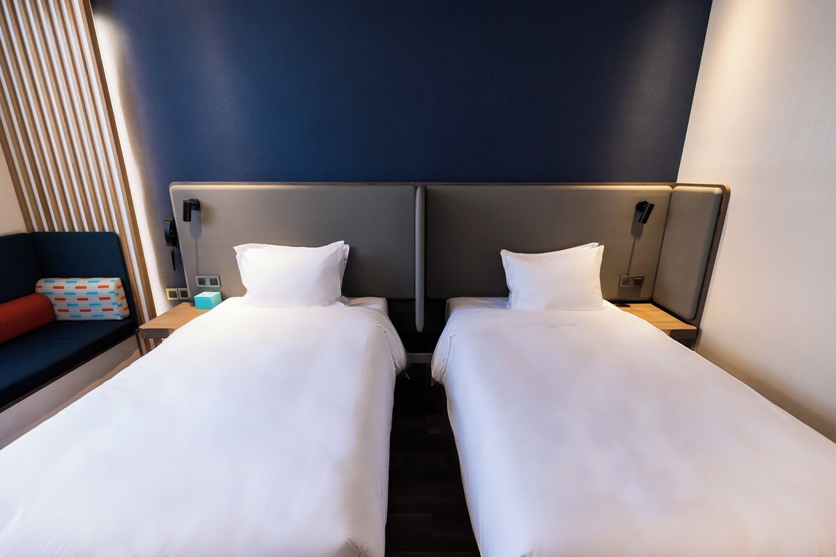 A hotel room with two single beds, a desk with a chair, and a window with blinds.