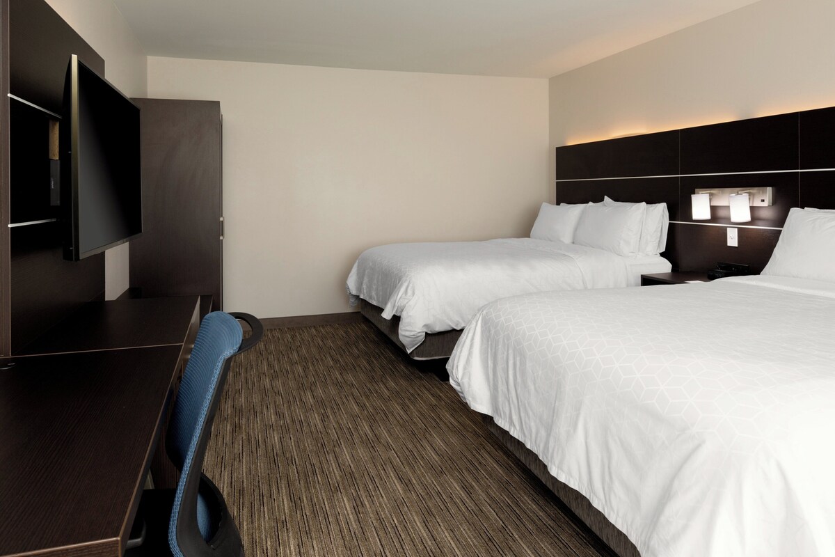 A hotel room with two beds, a desk, a TV, and a headboard with lights.