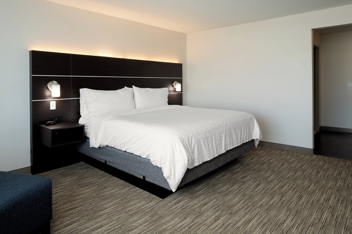 A modern hotel room with a large bed, dark headboard, bedside lamps, and a dark carpet.