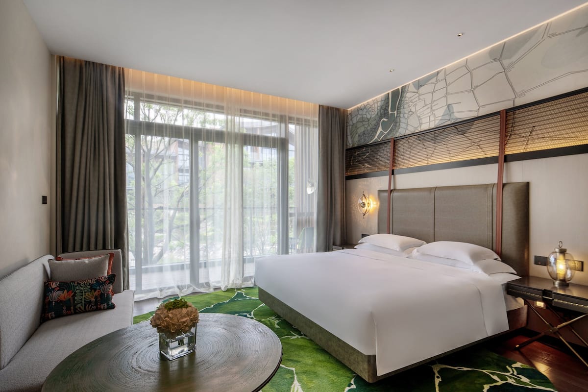 Una habitación de hotel moderna con una cama grande, un sofá, una mesa redonda y vistas a zonas verdes por la ventana.