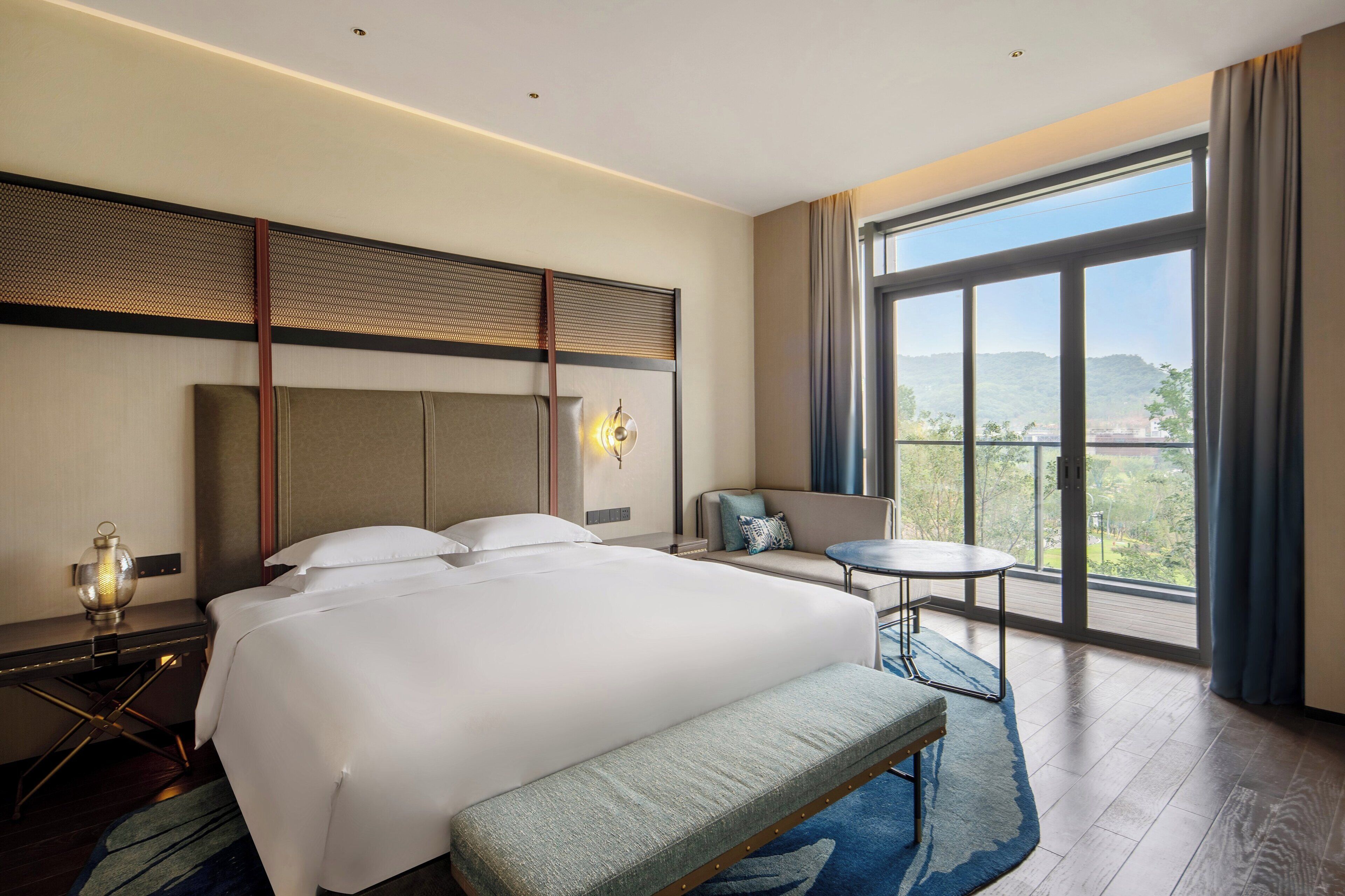 Habitación de hotel con una cama grande, un banco, una mesita y vistas al exterior.