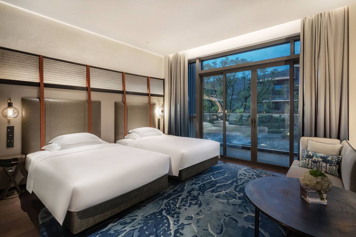 Habitación de hotel con dos camas, un ventanal amplio con vistas a árboles, una mesa redonda con un florero y una alfombra estampada.