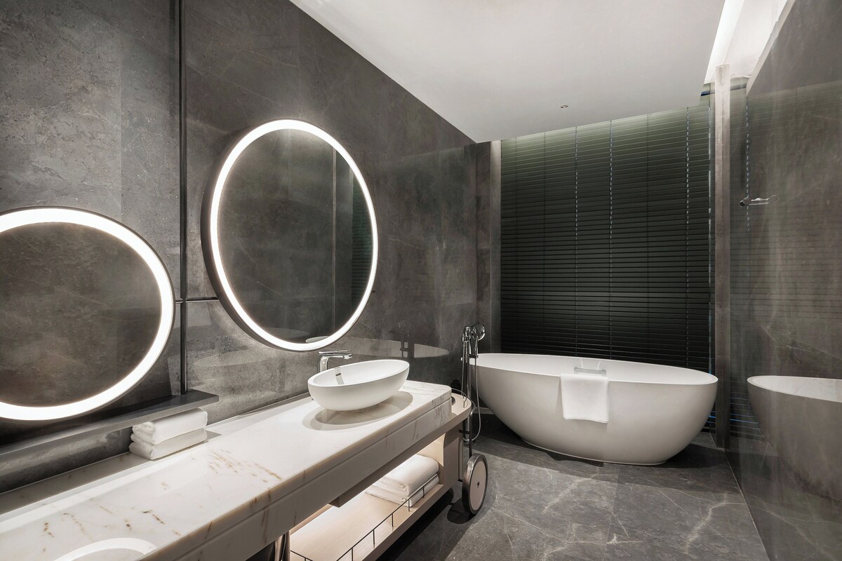 Un baño moderno con una bañera independiente grande, un espejo redondo con luz y una encimera de mármol.