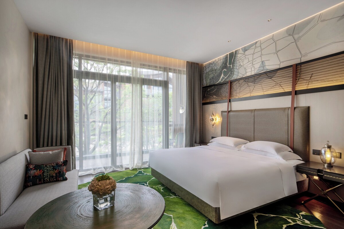 Una habitación de hotel moderna con una cama grande, un sofá, una mesa redonda y vistas a zonas verdes por la ventana.