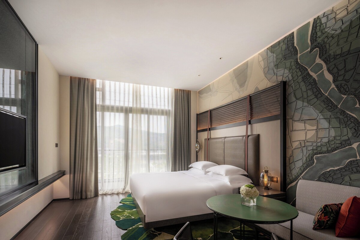 Habitación de hotel con una cama grande, una mesita pequeña, un sofá y un mural con un paisaje urbano en la pared.