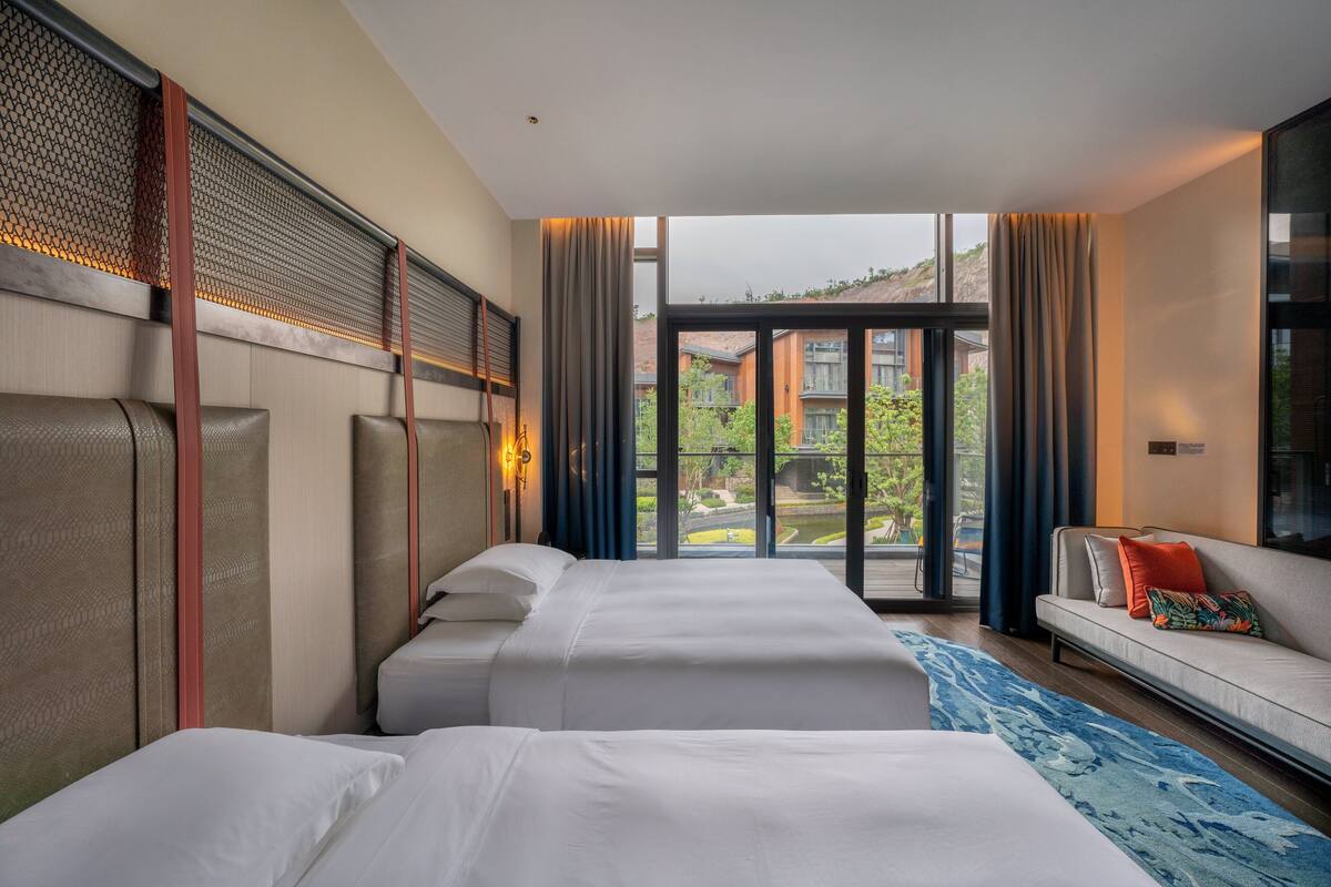 Habitación de hotel con dos camas, un sofá y un ventanal con vistas a edificios y vegetación.