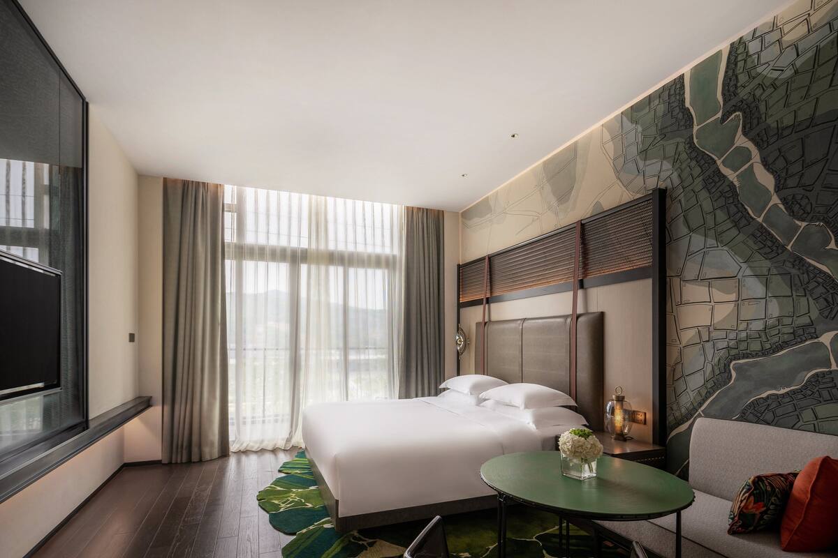 Habitación de hotel con una cama grande, una mesita pequeña, un sofá y un mural con un paisaje urbano en la pared.
