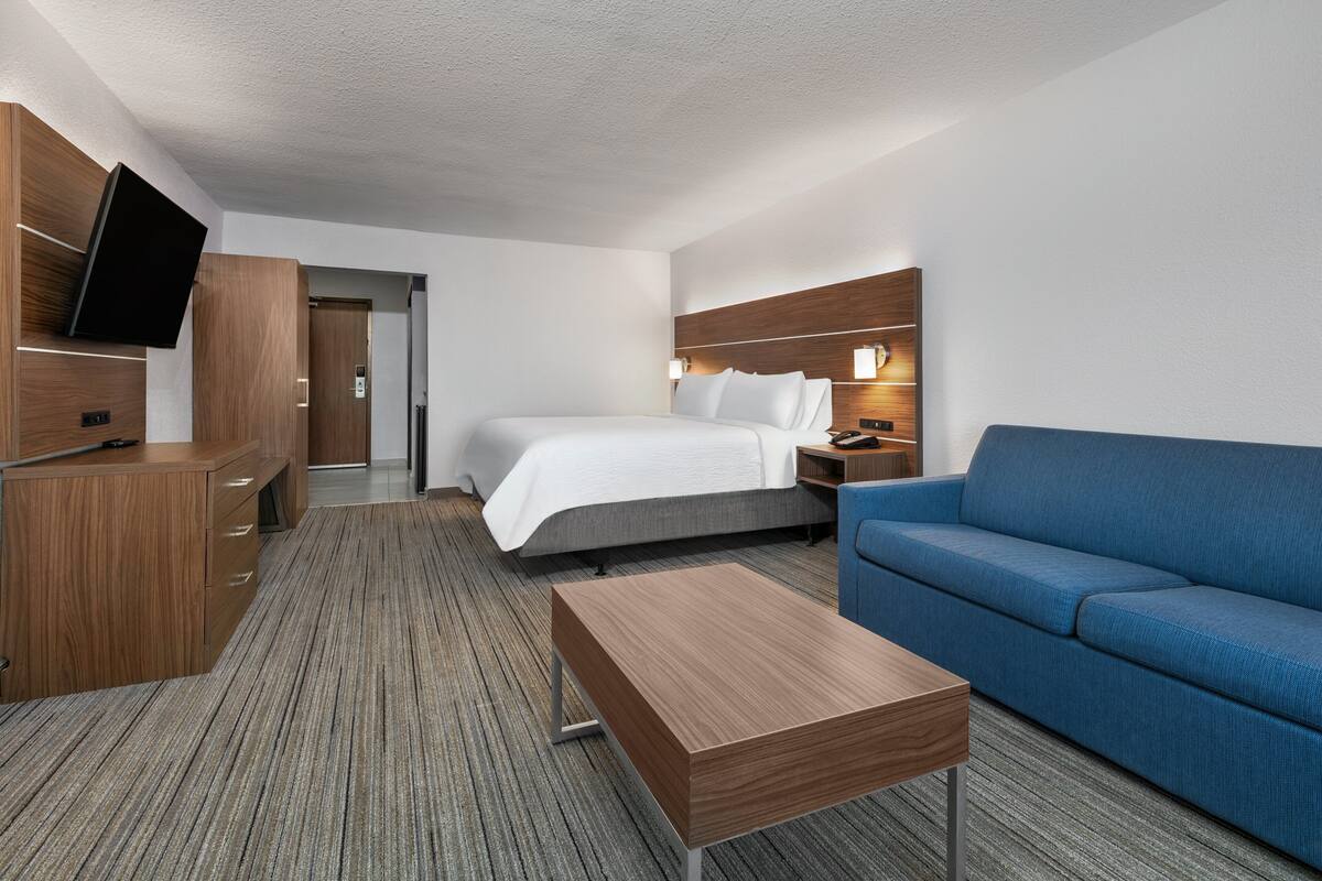 A hotel room with a bed, a blue sofa, a wooden desk, a flat-screen TV, and a nightstand with lamps.