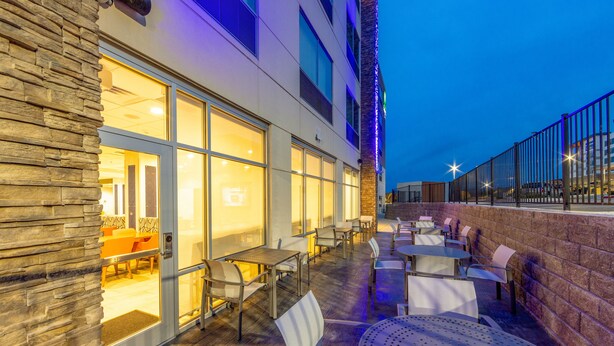 An outdoor patio area with tables and chairs, a stone wall, and a building with large windows. - Holiday Inn Express & Suites Harrisonburg University Area by IHG (Harrisonburg)