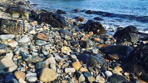 A rocky shoreline with a variety of stones and pebbles, a sandy beach, and a forested cliff in the background. - Sunrise Suite, near Rockland Harbor, walk to DT (Rockland)