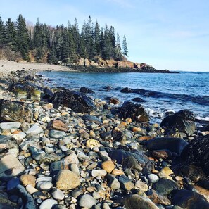 A rocky shoreline with a variety of stones and pebbles, a sandy beach, and a forested cliff in the background.