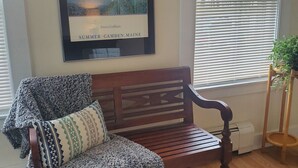 Un banc de fusta amb una manta decorativa i coixins, un quadre emmarcat de velers i una planta en test amb vistes a una finestra amb persianes.