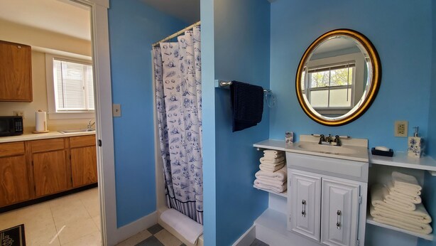 A bathroom with a blue wall, a shower curtain with a pattern, a sink with a mirror, and towels on a shelf. - Sunrise Suite, near Rockland Harbor, walk to DT (Rockland)