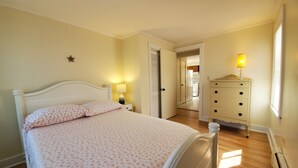 Un dormitori amb llit, taulella de nit, làmpada, prestatge i una porta que dóna a una altra habitació.