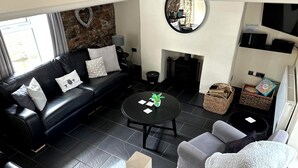 Ein modernes Wohnzimmer mit einer schwarzen Ledersofa, einem runden Couchtisch und einem grauen Sessel. Im Hintergrund ist ein Kamin zu sehen, und auf dem Boden stehen Korbwäschebehälter.