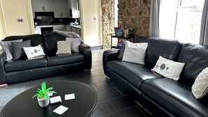Ein modernes Wohnzimmer mit zwei schwarzen Ledersofas, einem runden schwarzen Couchtisch mit Pflanze und Spielkarten, und einer durch einen offenen Durchgang sichtbaren Küche.