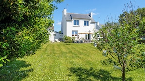 Une maison à deux étages avec une pelouse bien entretenue, une terrasse dotée de mobilier d’extérieur et un ciel d’un bleu éclatant.