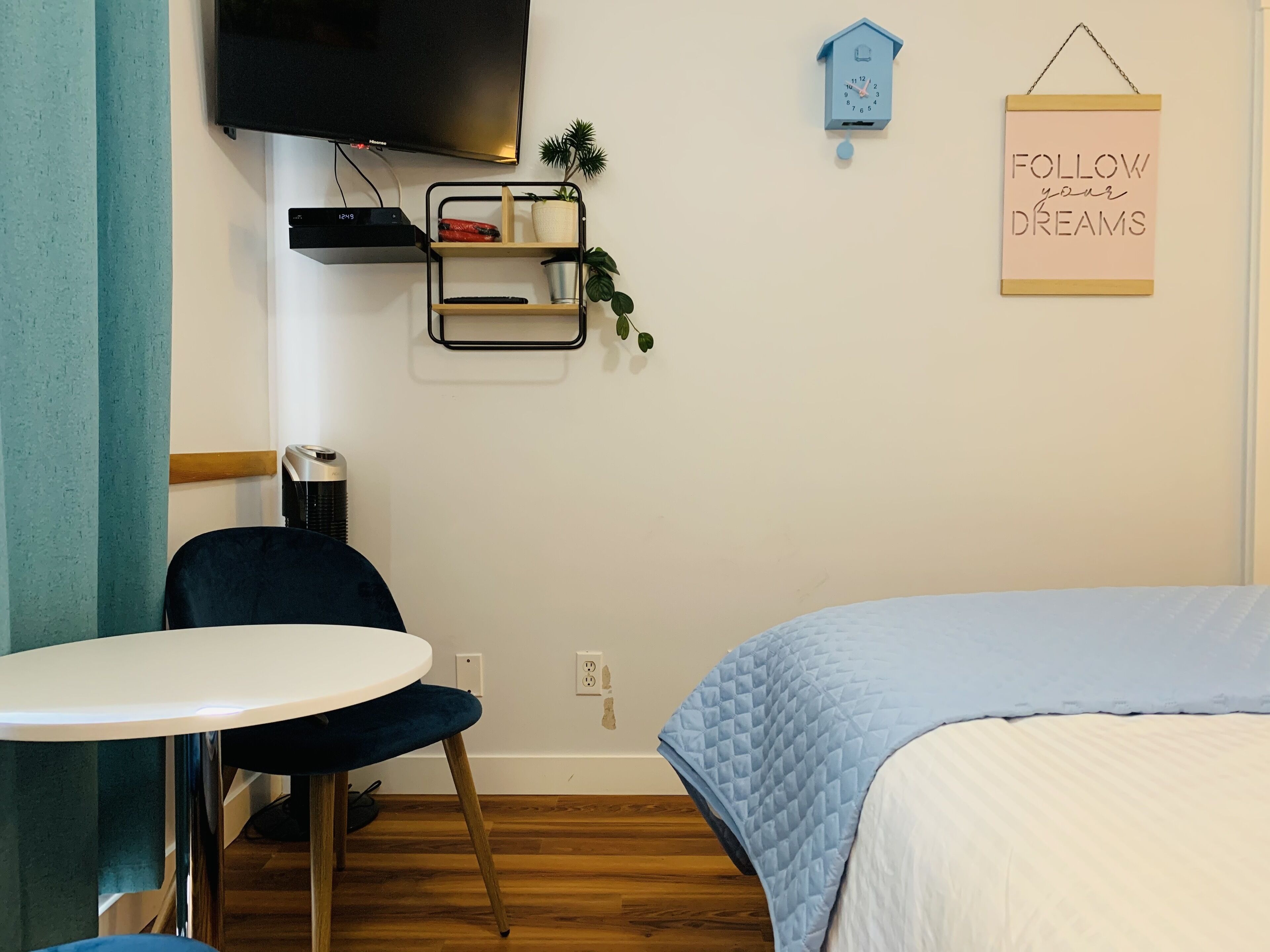 A neatly arranged room with a bed, a table, a chair, a TV, a clock, and a motivational sign.