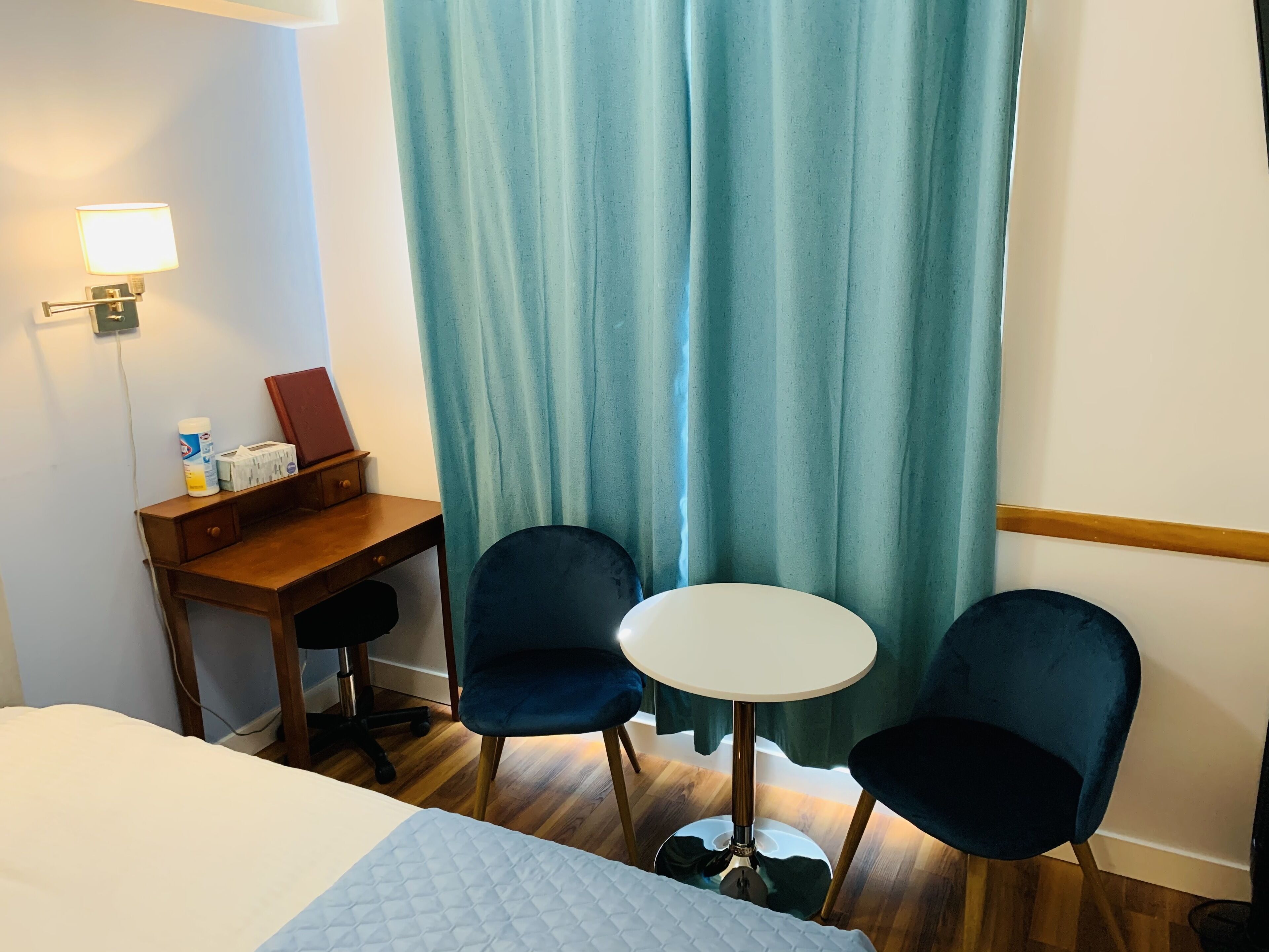 A hotel room with a bed, a desk, two chairs, a small table, a lamp, and a curtain.