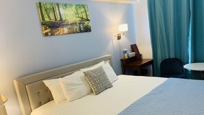 A hotel room with a bed, a desk, a chair, a painting of a forest, and a window with curtains. - A Small Hotel Room for Mountain Lovers (Canmore)
