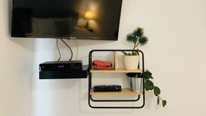 A flat-screen TV mounted on a wall with a shelf below it containing a plant, a basket, and a remote control. - A Small Hotel Room for Mountain Lovers (Canmore)