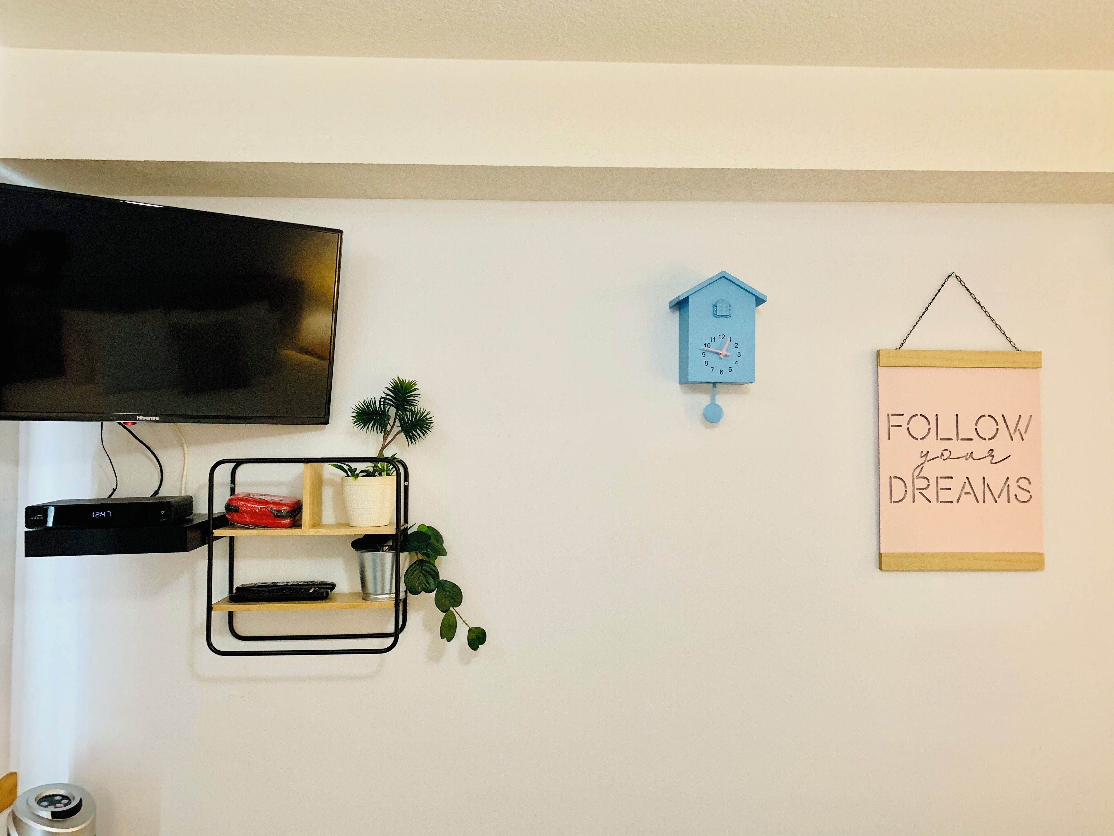Een tv aan de muur, een blauwe kuihorloge, een plank met planten en een rood voorwerp, en een motiverende poster.