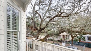 A balcony with a white railing, overlooking a street with parked cars and a building with a porch.
