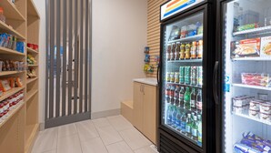 Une vitrine réfrigérée contenant diverses boissons et collations, placée à côté d’un meuble en bois et d’un présentoir vertical où sont exposés des produits emballés.