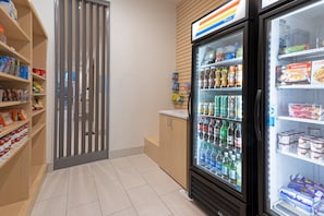 A refrigerated display case with various beverages and snacks, adjacent to a wooden cabinet and a vertical display of packaged goods. - Holiday Inn Express & Suites Firestone Longmont by IHG (Longmont)