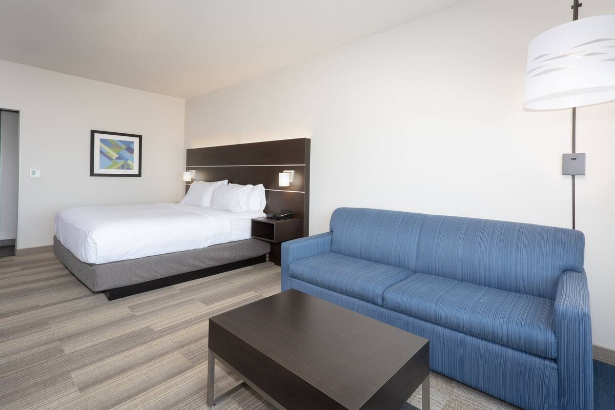A hotel room with a bed, a blue sofa, a coffee table, a bedside table with a lamp, and a framed picture on the wall.