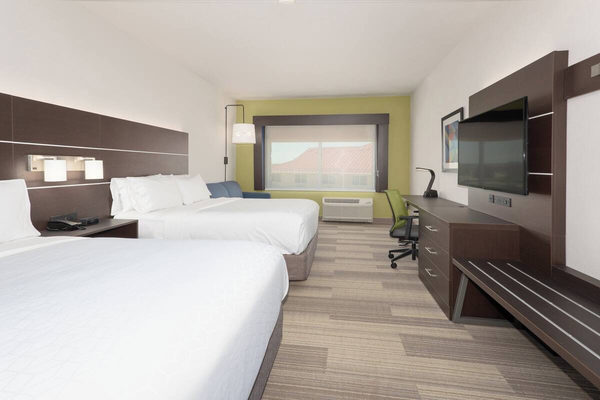A hotel room with two beds, a desk, a chair, a TV, and a window with a view of buildings.