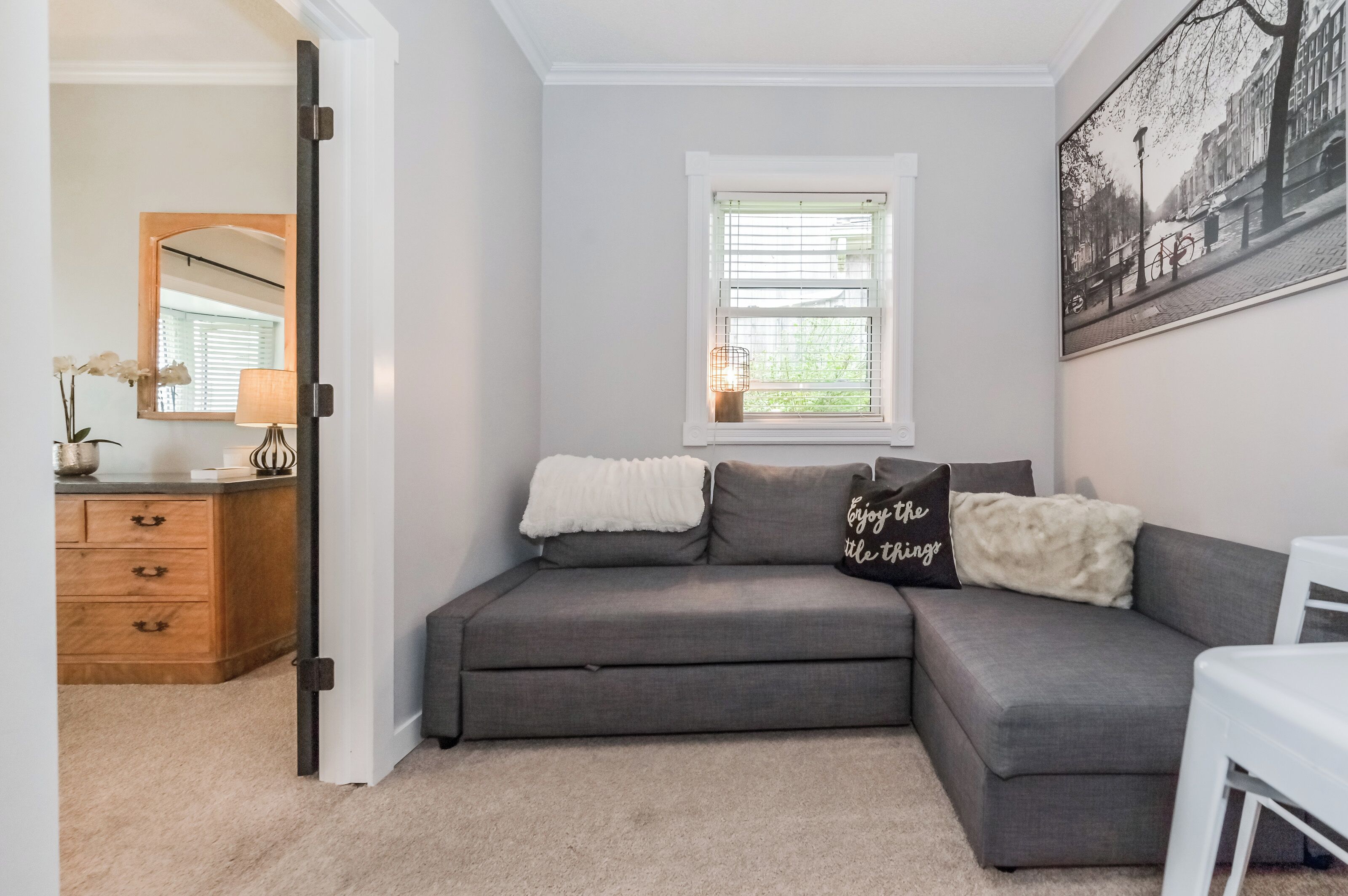 A grey sectional sofa with a white throw blanket and a pillow with 'Enjoy the title' text, a window with blinds, a framed picture on the wall, and a wooden dresser with drawers.