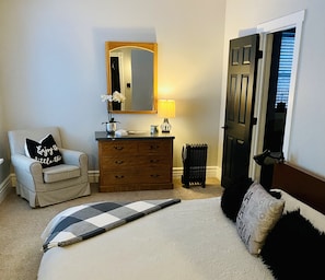 A bedroom with a bed, a chair, a dresser, a mirror, and a door. - The VICTORIA: Petit Appartment (Elora)