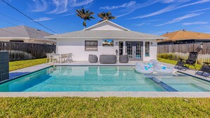 A residential swimming pool with lounge chairs and inflatable floats, surrounded by a wooden fence and palm trees.
