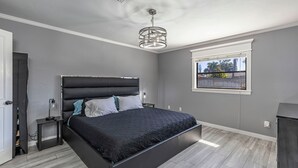 A bedroom with a large bed, a bedside table, a nightstand, a ceiling light fixture, and a window with a view of palm trees.
