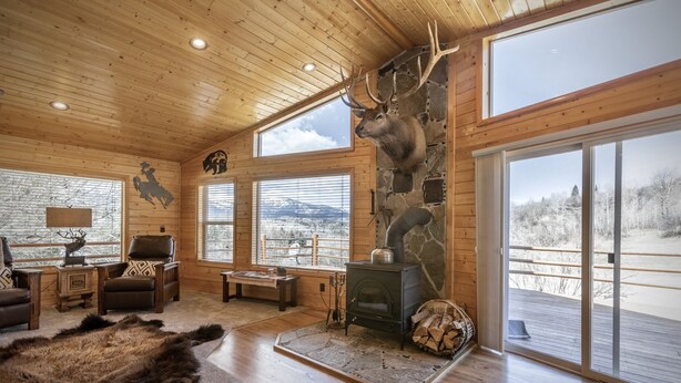 A log cabin interior with a stone wall, wooden ceiling, large windows, a fireplace, and a mounted elk head.