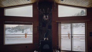 A log cabin with a stone fireplace, wooden floors, and large windows offering views of a snowy landscape.