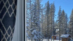 Ikkunasta avautuu näkymä lumiseen metsään, jossa on korkeita puita ja näkyvillä on myös puinen rakennelma.
