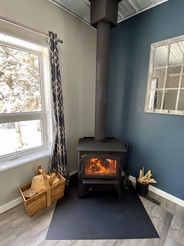 A room with a wood-burning stove, a window with a view of trees, and a wooden box of firewood.