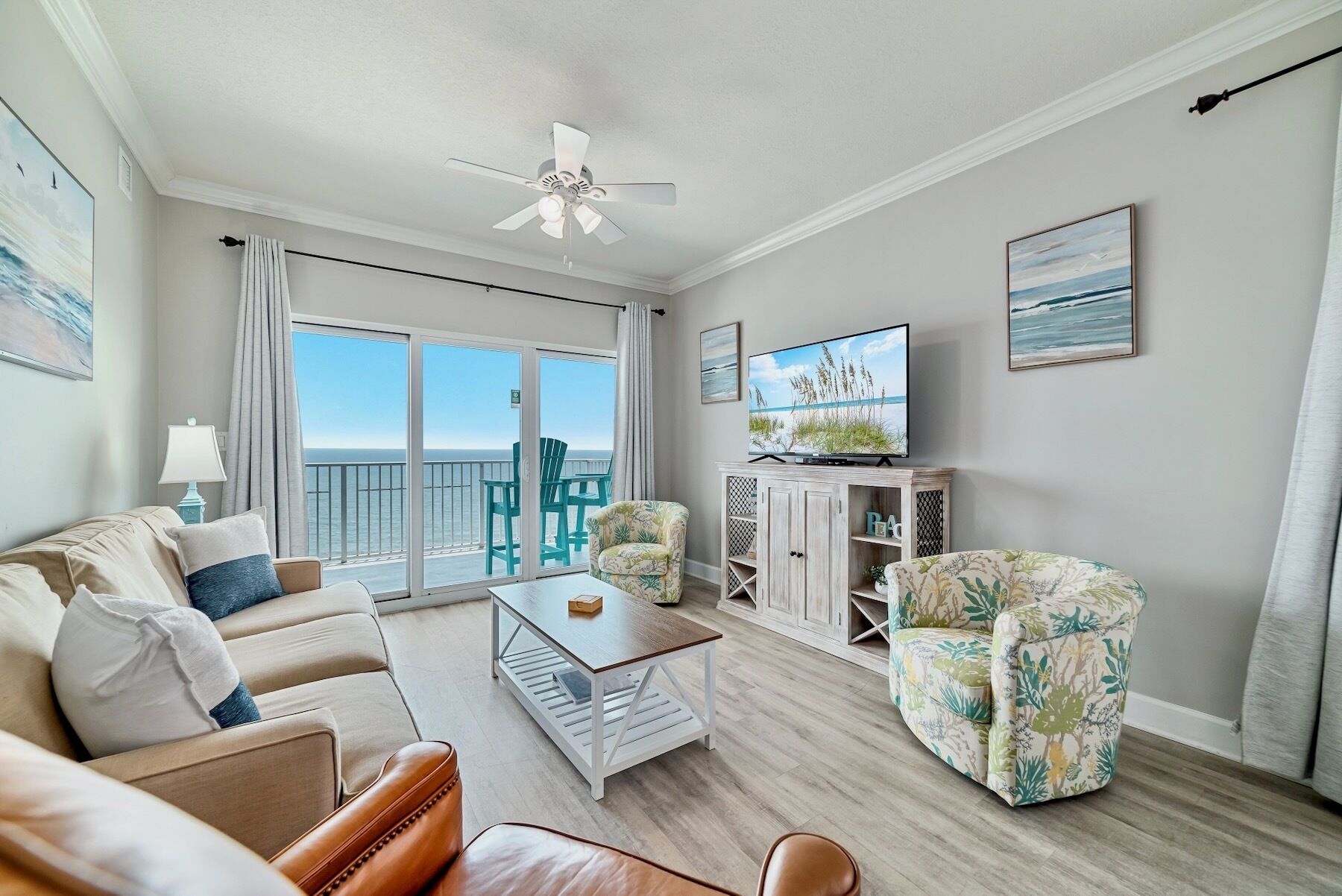 A living room with a large window, a ceiling fan, a television, and a view of the ocean.