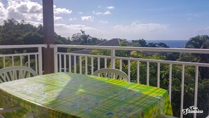 A balcony with a table covered in a green and yellow checkered tablecloth, white chairs, and a view of the ocean and hills. - Location Appartement / Gite à Deshaies, Calme de la Montagne et vue sur mer (Deshaies)