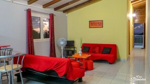 A room with a red sofa, a small dining table, a fan, and a window with red curtains. - Location Appartement / Gite à Deshaies, Calme de la Montagne et vue sur mer (Deshaies)