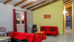 Une pièce avec un canapé rouge, une petite table à manger, un ventilateur et une fenêtre avec des rideaux rouges.