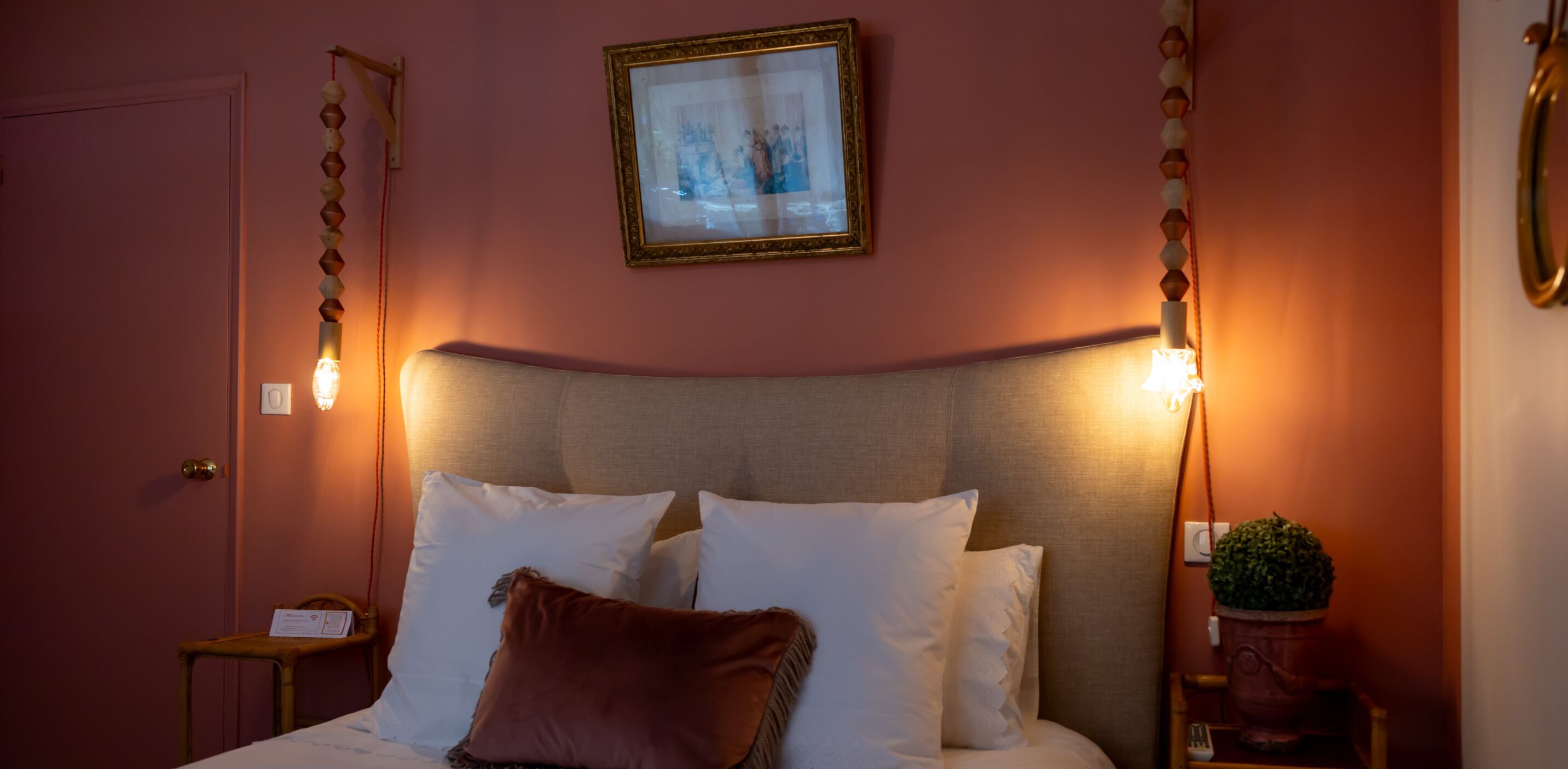 A neatly made bed with white and brown pillows, a framed picture on the wall, and a potted plant on a side table.