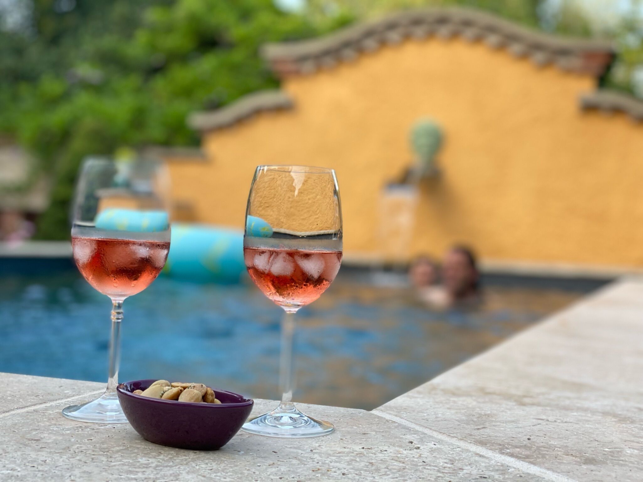 To glas rosa drik med isterninger på et bord ved poolen, en skål med nødder og en pool i baggrunden.